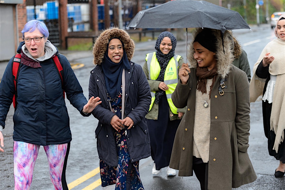 A group of five women walk together outdoors on wet pavement on an overcast day. In the foreground, a person on the left with purple hair and glasses, wearing a blue jacket and colorful leggings, gestures while talking. Next to them, a woman in a blue parka with a fur-trimmed hood and a hijab smiles. On the right, another woman in a green coat and black head wrap smiles as she holds a black umbrella over herself and the woman next to her. In the background, two other women, one wearing a high-visibility vest and a hijab, follow and smile.