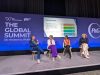 Four women are seated on stage in chairs during a panel discussion at The Global Summit on Women & Sport. Behind the speakers, a large blue backdrop features logos for IWG Women & Sport, Sport + Recreation Alliance, and P&G. A central presentation screen titled "Get on Board" outlines four key steps related to the Brighton Plus Helsinki Declaration. From left to right on stage, a speaker wearing a blue suit sits cross-legged while speaking into a microphone, followed by a speaker in a white jacket over a long purple dress who listens attentively. Beside her, a speaker wears an orange sleeveless top with black trousers, and on the far right, a speaker is dressed in a dark jacket with light-colored trousers.