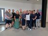A group of twelve people stand together indoors posing for a group photograph in front of a white wall. In the center, a woman in a navy polka-dot blouse and dark skirt and a woman in a navy blazer and trousers jointly hold a framed document titled "Brighton plus Helsinki Declaration on Women and Sport." The surrounding group members are dressed in a mix of smart-casual and athletic attire, including t-shirts, polo shirts, skirts, and trousers. On the far right, a Liverpool John Moores University banner stands next to a large black structural pillar.