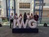 A group photo of seven people standing in a line behind a large 3D white display spelling out "IWG" on a dark blue base. The group is posed along an indoor balcony railing inside a spacious, multi-story building atrium with large windows and blue structural pillars. A dark blue banner hanging on the right side reads "THE GLOBAL SUMMIT ON WOMEN".
