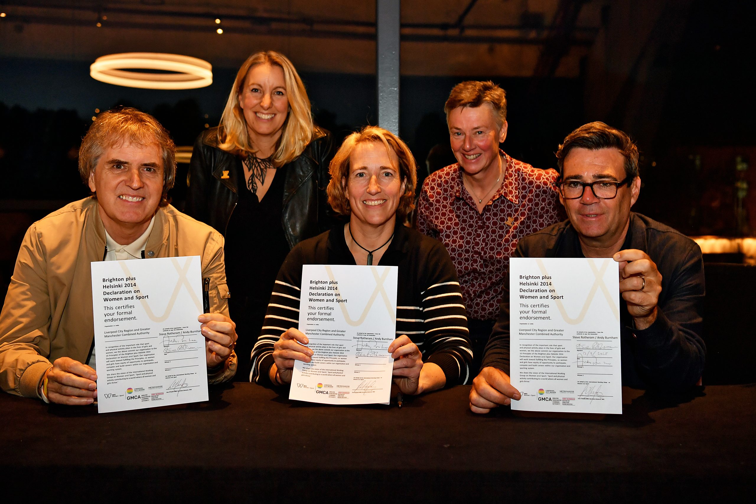 Five people smile around a table at an indoor event, with three seated individuals holding up signed copies of the Brighton plus Helsinki 2014 Declaration on Women and Sport.