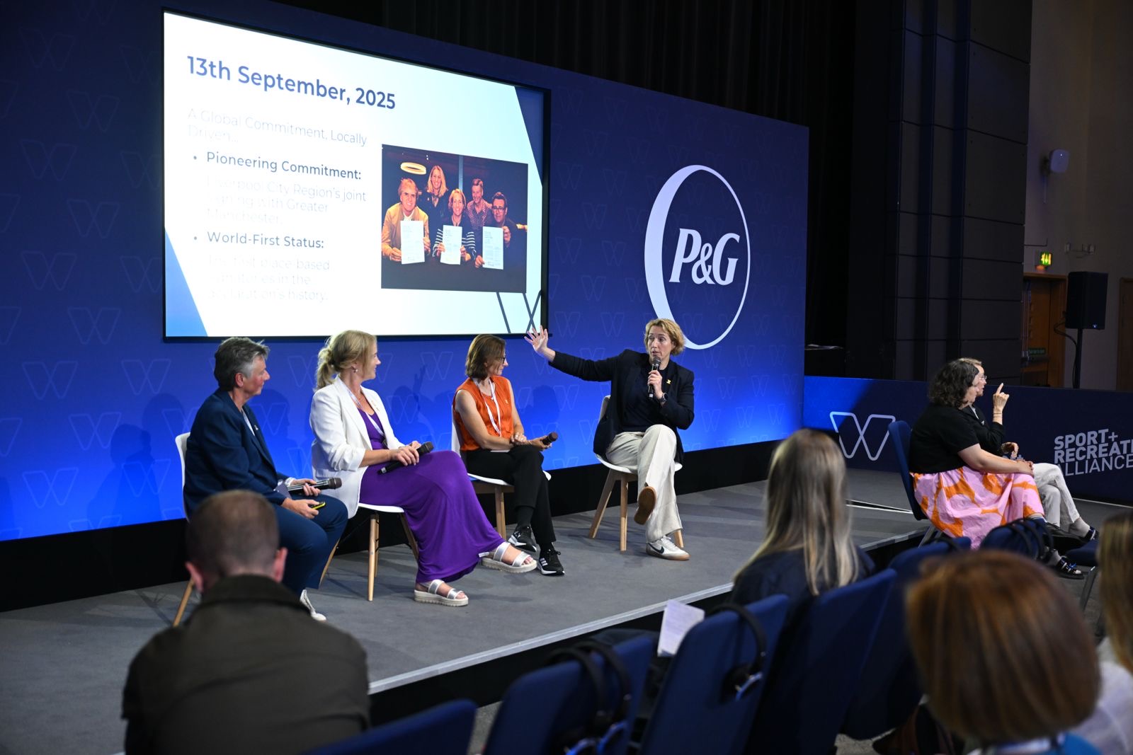 A panel of four speakers and a BSL interpreter present on stage at a conference. One speaker gestures into a microphone in front of a large screen displaying a photo of a signing ceremony and text dated 13th September, 2025.