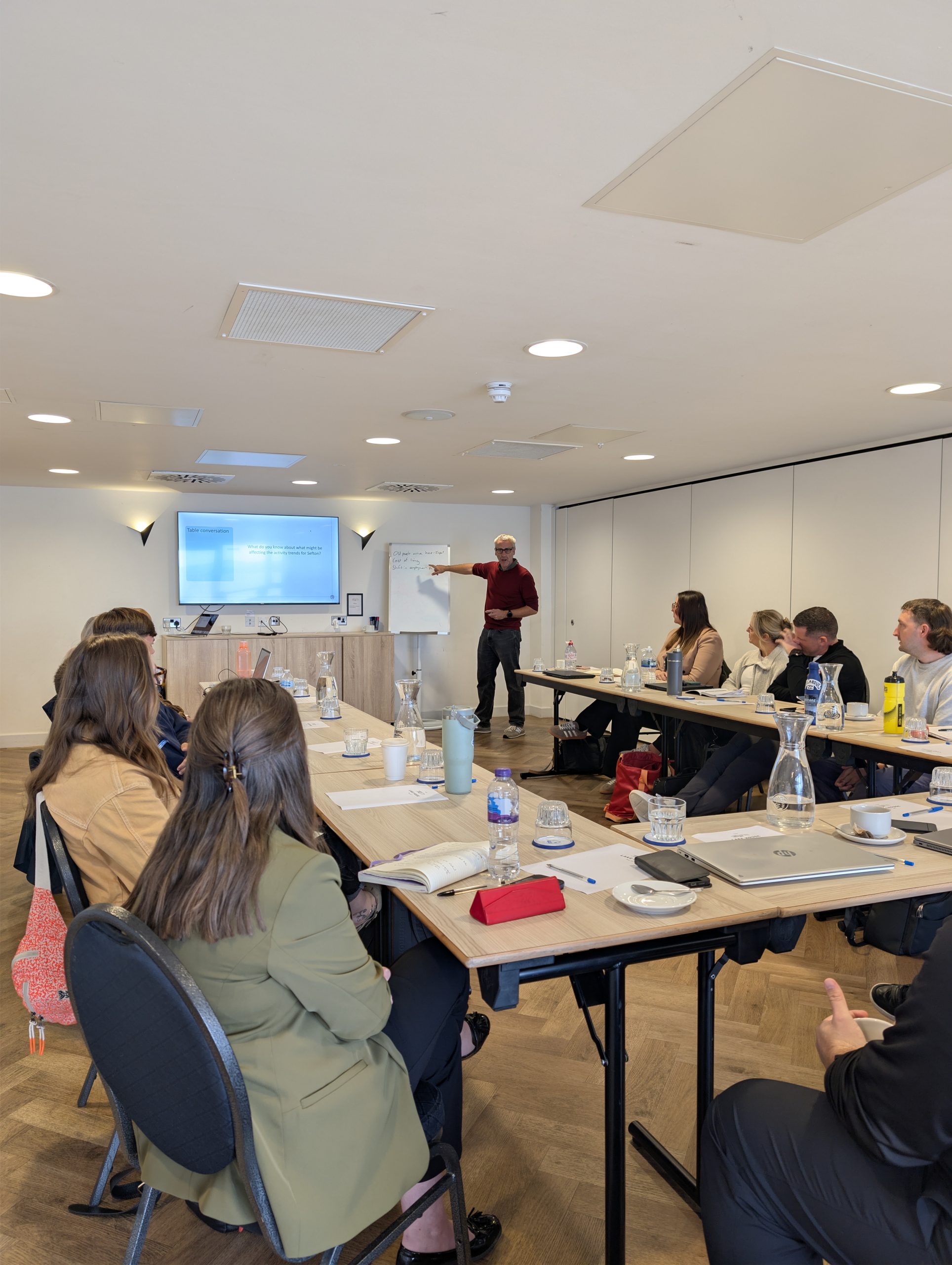 A man wearing glasses and a maroon sweater stands at the front of a brightly lit meeting room, pointing toward notes written on a whiteboard next to a wall-mounted TV screen displaying a presentation slide titled "Table conversation." Grouped around tables arranged in a U-shape, several attendees sit listening to the presentation. The tables are set with laptops, notebooks, water carafes, and glasses, while two women in the foreground have their backs turned slightly toward the camera as they face the speaker.